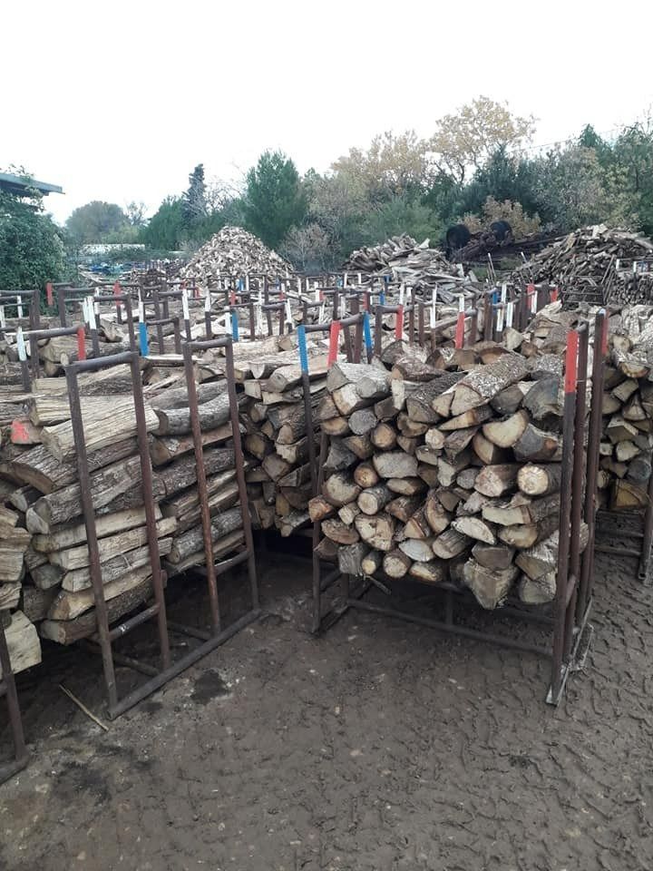 Des piles de bois de chauffage coupé sur des étagères métalliques dans une cour extérieure, avec des arbres en arrière-plan.