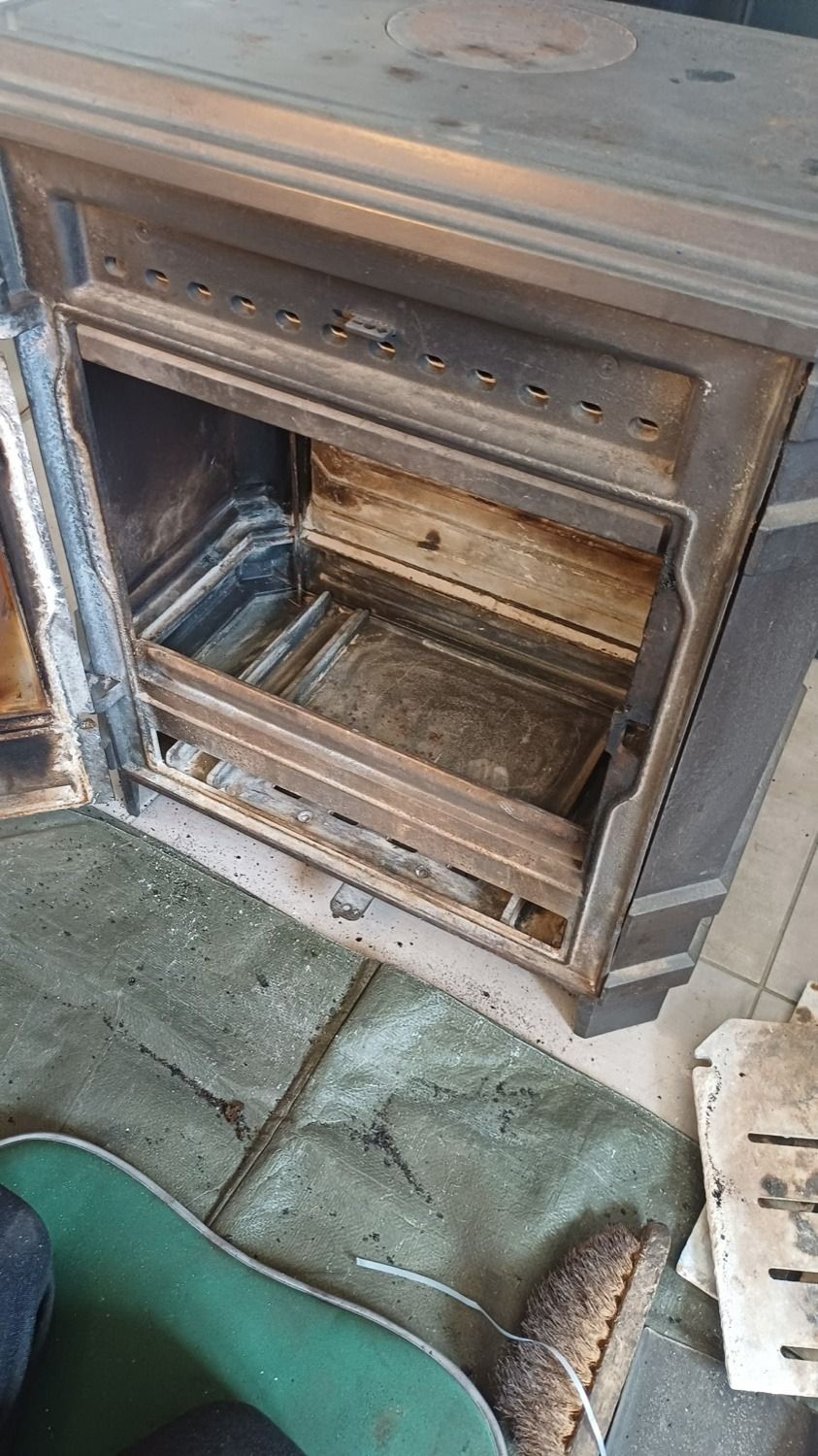 Poêle à bois rouillé, porte ouverte, laissant apparaître un foyer vide et un tapis de sol usé à proximité.