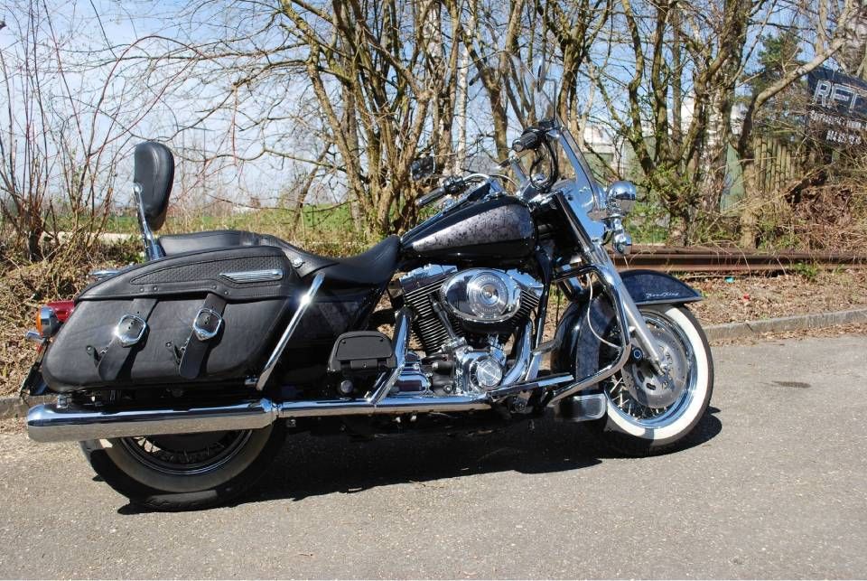 Schwarze Harley-Davidson mit Satteltaschen und Windschutzscheibe, geparkt in der Nähe von Bahngleisen und Bäumen.