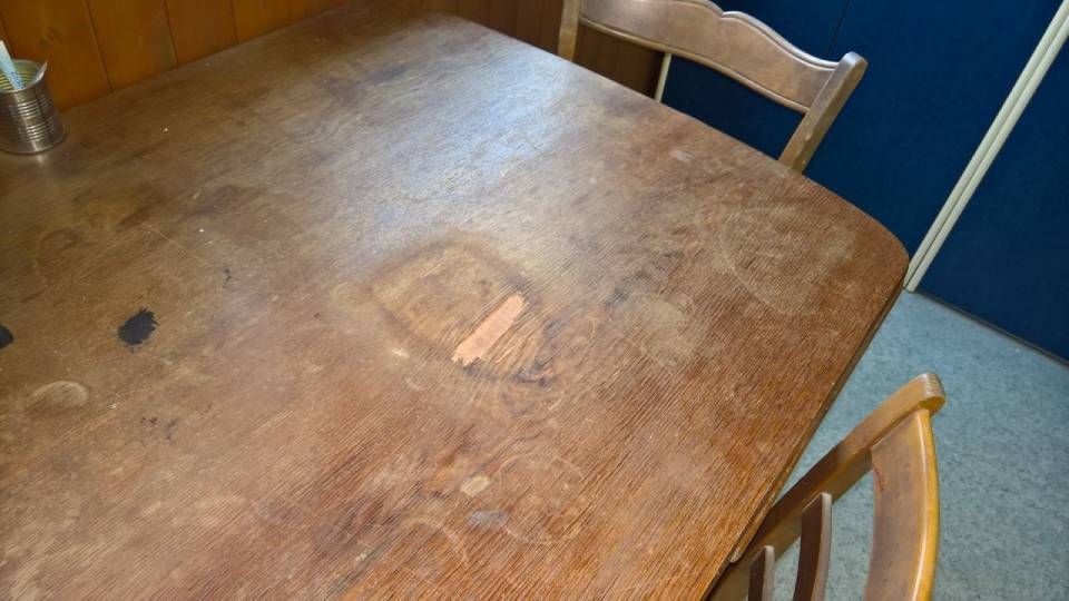 Holztisch mit sichtbaren Gebrauchsspuren, zwei Stühle teilweise sichtbar.