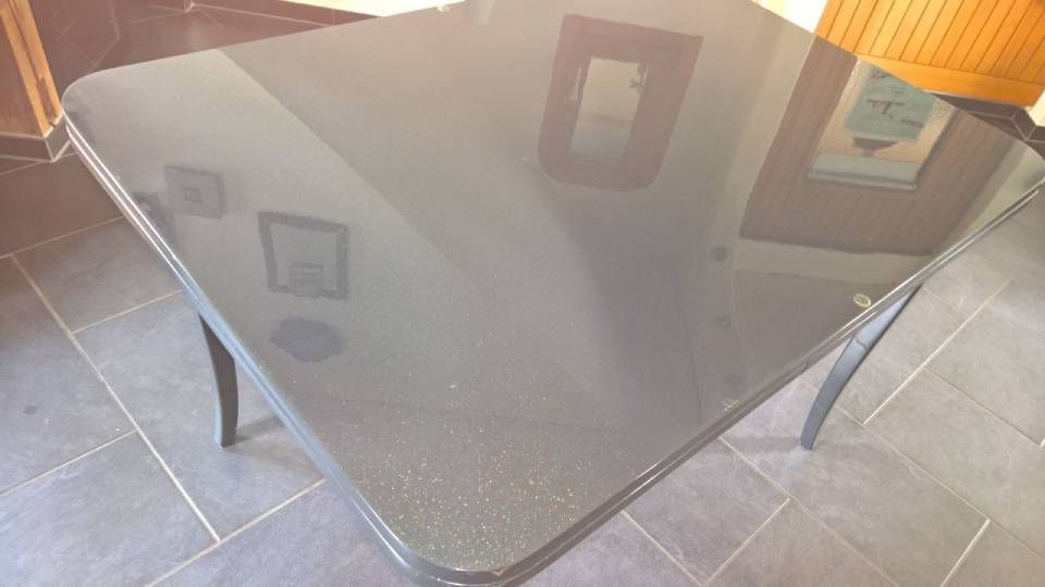 Ein glänzender, schwarzer quadratischer Tisch mit geschwungenen Beinen steht auf einem Fliesenboden und spiegelt den Raum wider.