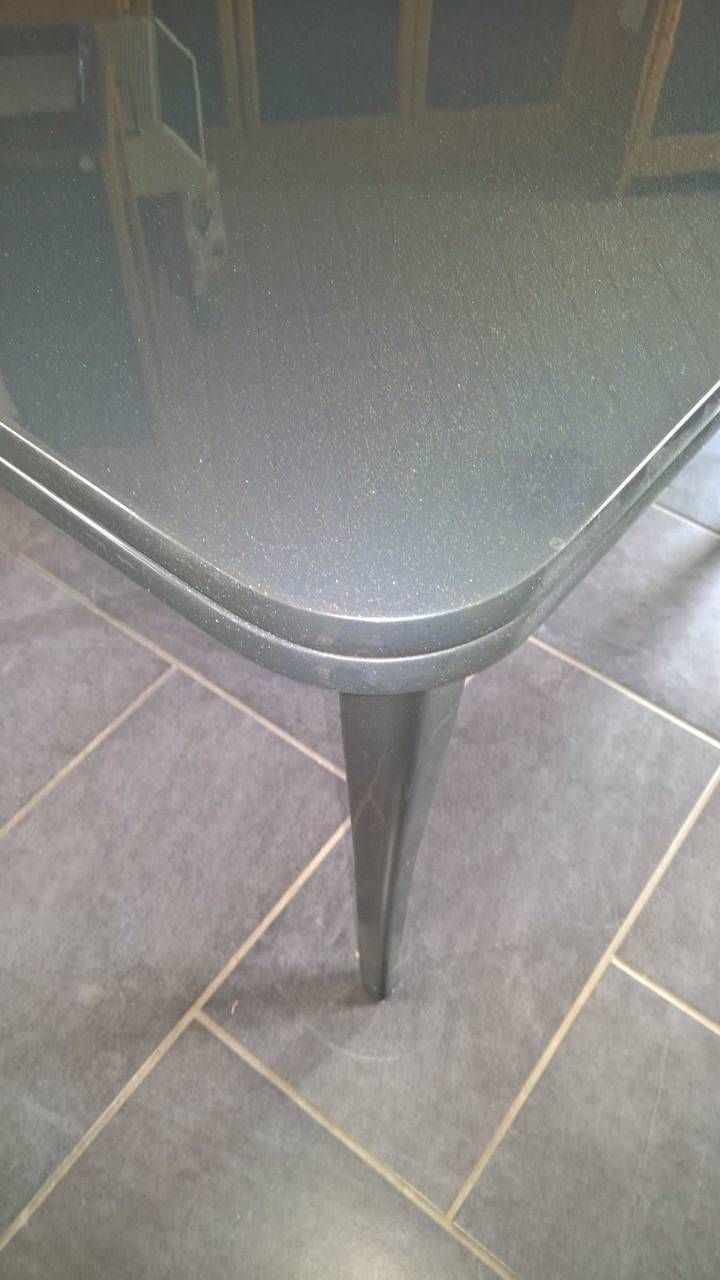 Dunkelgrauer Tisch mit abgerundeten Kanten, der auf einem gefliesten Boden steht. Ein Tischbein ist sichtbar.