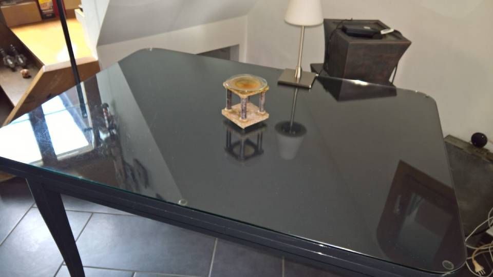 Ein dunkler Schreibtisch mit Glasplatte und darauf befindlichen Gegenständen in einem Raum mit Lampe und Fliesenboden.