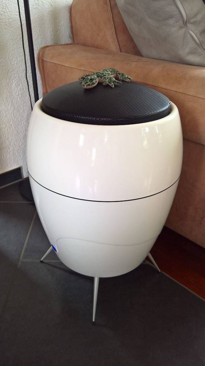 Weisser, eiförmiger Kompostbehälter mit schwarzem Gitterdeckel und Metallbeinen, drinnen in der Nähe eines Sofas.