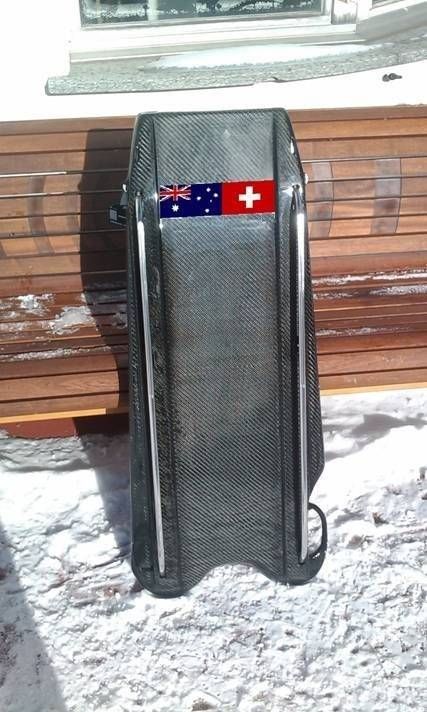 Schlitten mit Flaggen von Australien und der Schweiz auf Schnee vor einer Holzmauer.