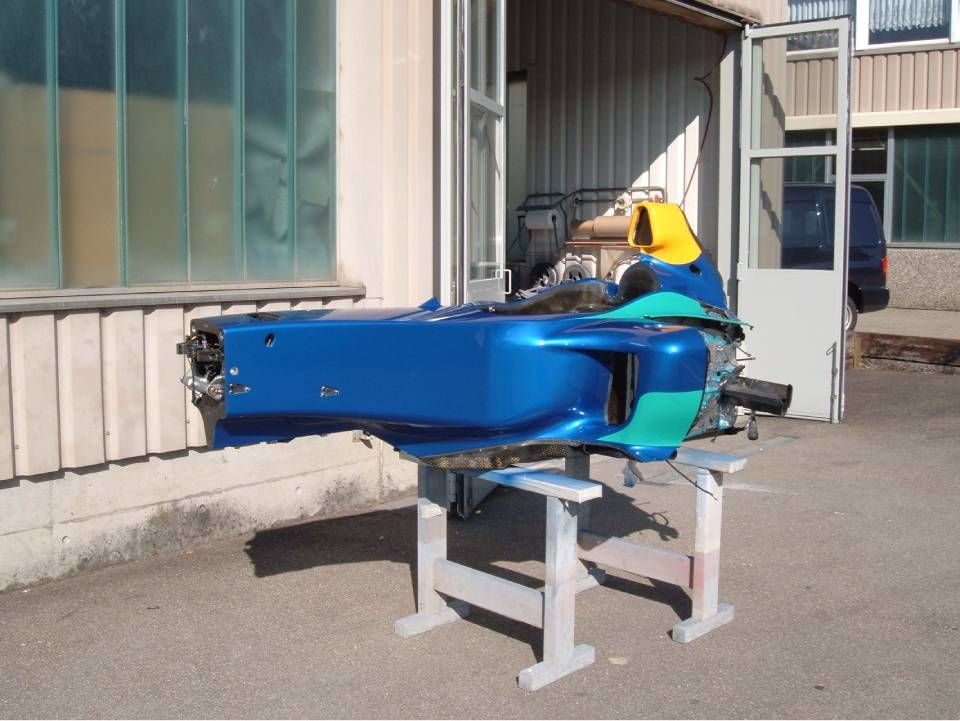 Blaue und grüne Rennwagenchassis auf einem weissen Ständer im Freien neben einem Gebäude.