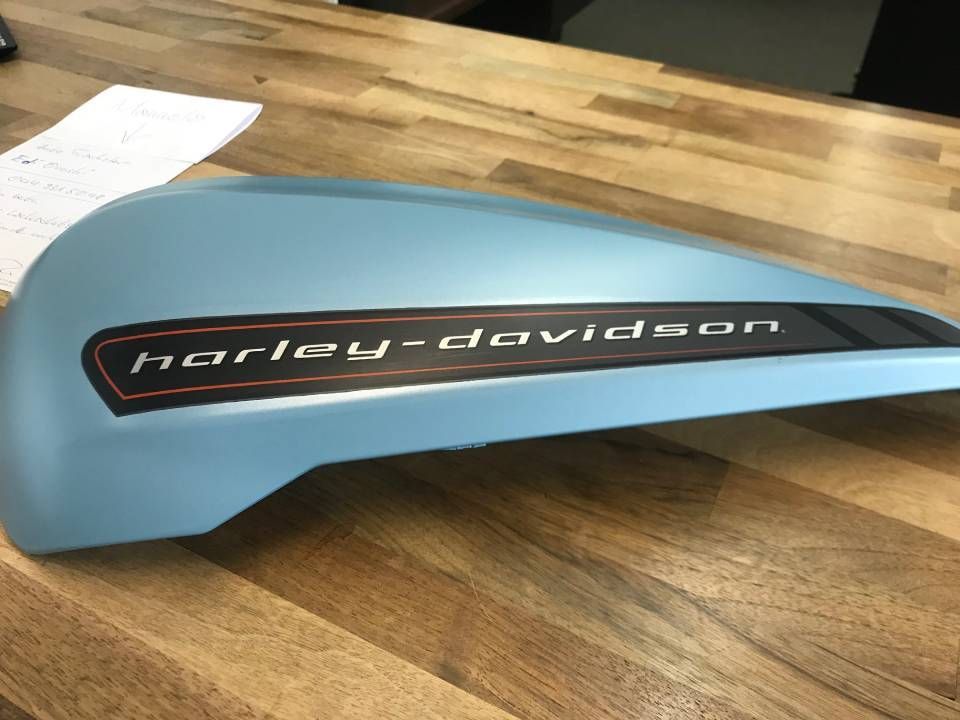 Hellblauer Benzintank eines Harley-Davidson-Motorrads mit schwarzem und orangefarbenem Logo auf einem Holztisch.