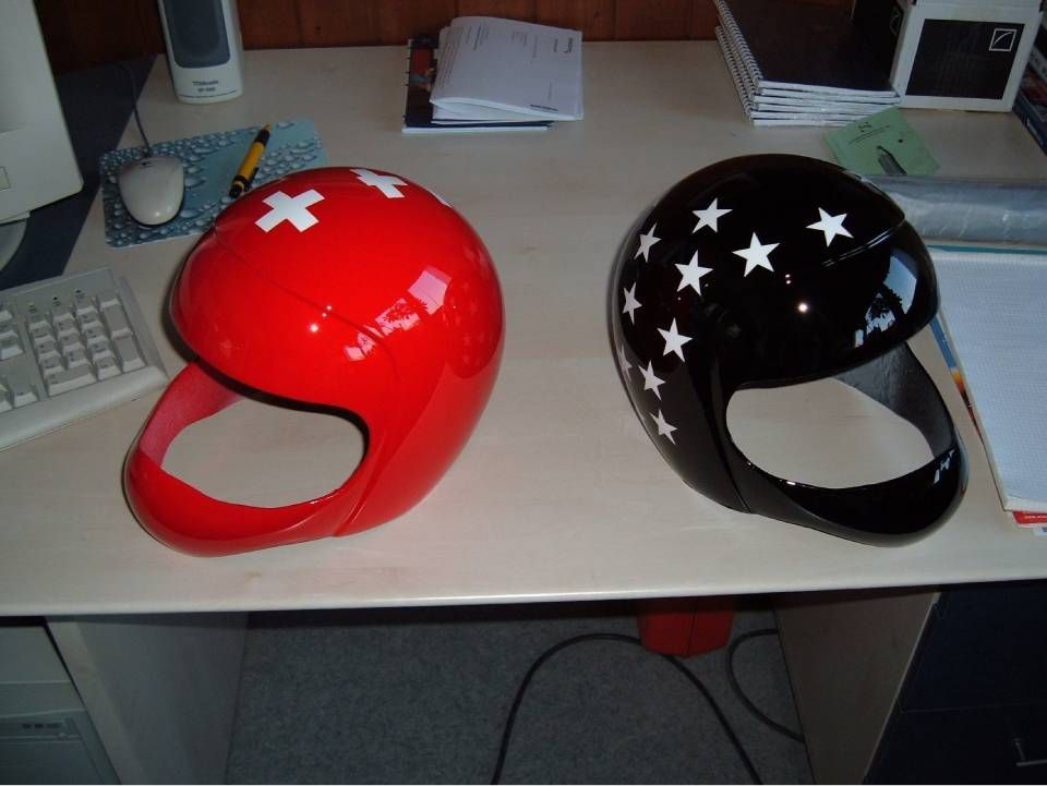 Zwei glänzende Rennhelme: ein roter mit weissen Kreuzen und ein schwarzer mit weissen Sternen, auf einem Schreibtisch.