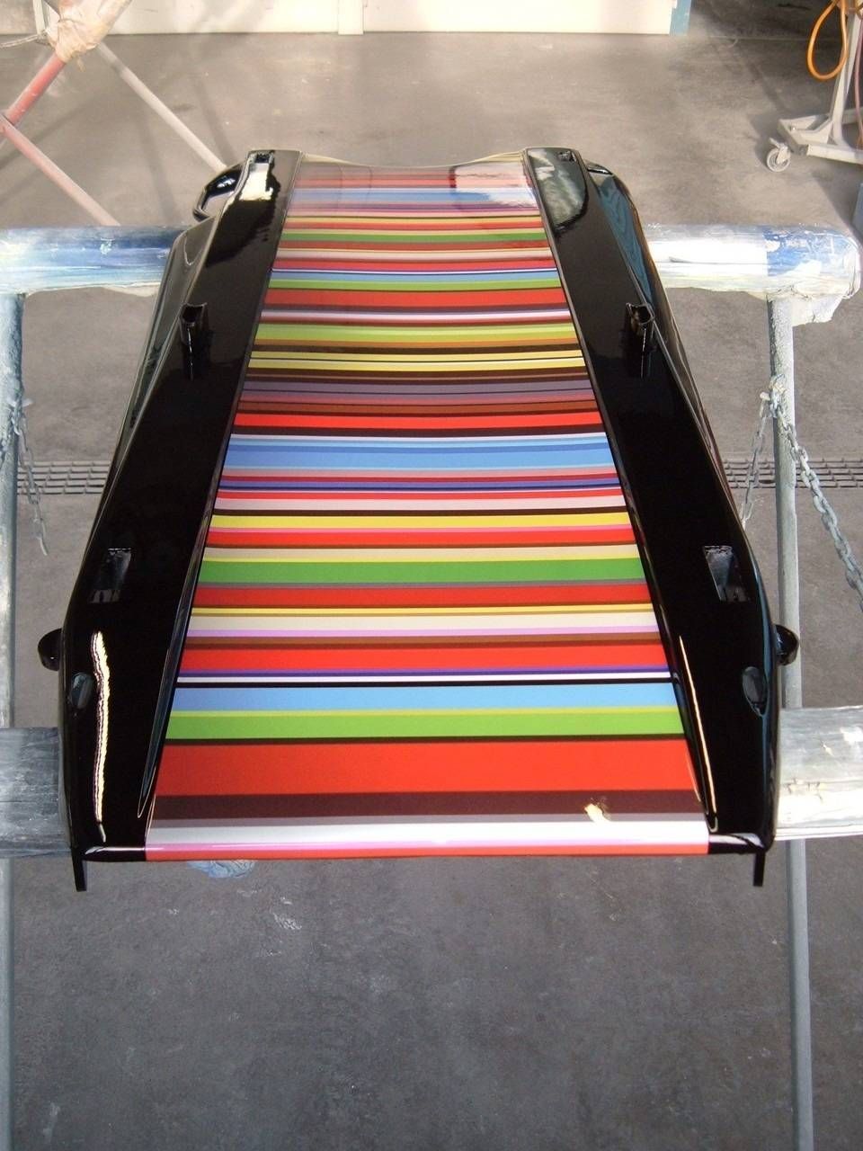 Schwarzes Metallobjekt mit regenbogenfarben gestreifter Innenseite, ruhend auf einem blauen Sockel, in einer hellen Umgebung.