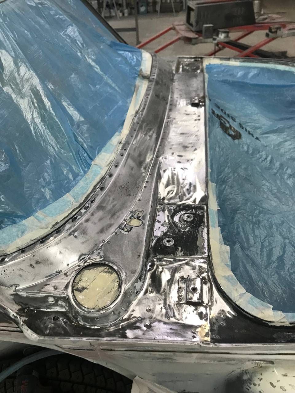 Die Karosserie wird repariert; blankes Metall und Abklebeband sind sichtbar. Blaue Plastikfolie schützt die angrenzenden Bereiche.