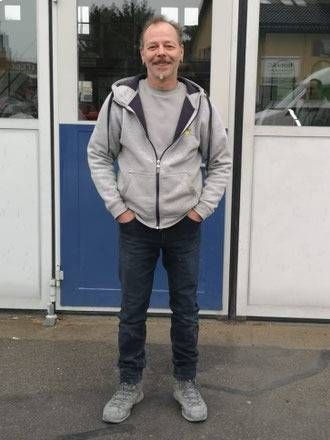 Mann in grauem Kapuzenpulli und Jeans, Hände in den Taschen, steht vor einer blauen Tür und Fenstern mit weissen Rahmen.