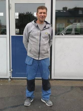 Ein Mann in Arbeitskleidung steht vor einem Gebäude. Er trägt eine graue Kapuzenjacke und eine blaue Arbeitshose.