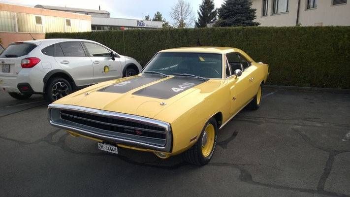 Ein gelber Dodge Charger Muscle-Car aus dem Jahr 1970 parkt neben einem silbernen SUV auf einem gepflasterten Parkplatz.