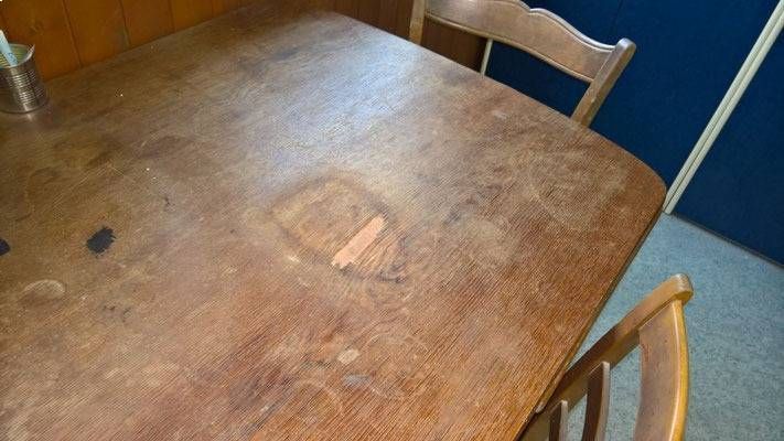 Ein Holztisch mit abgenutzter Oberfläche, zwei Stühle sind zu sehen, im Hintergrund eine Wand.