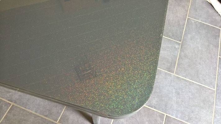 Nahaufnahme einer abgerundeten Ecke eines dunklen, gesprenkelten, grün-grauen Tisches auf einem gefliesten Boden.