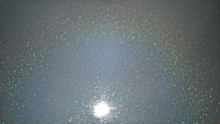 Helle, weisse Lichtquelle, umgeben von gesprenkelten, irisierenden Farben auf grauem Hintergrund.