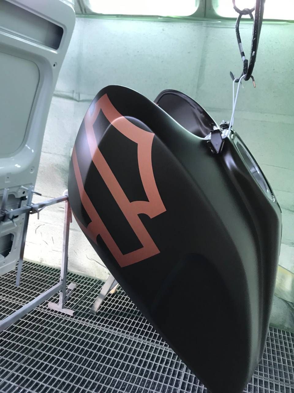 Motorradtank schwarz lackiert mit kupferfarbenem Harley-Davidson-Logo. Hängt in einer Lackierkabine.