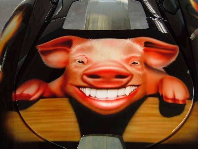 Ein lächelndes Schwein, mit Airbrush auf eine schwarze Motorhaube gesprüht, lehnt sich über einen Holzzaun.