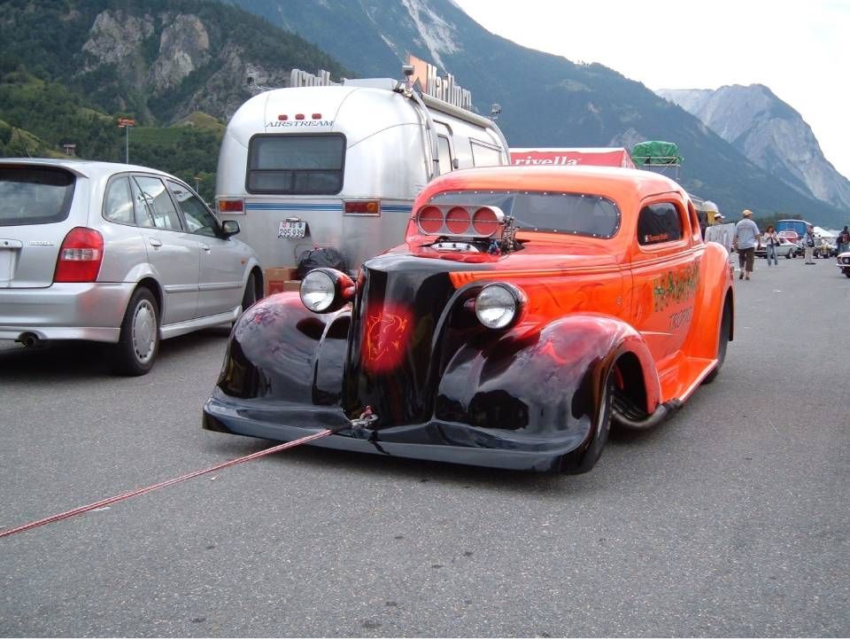Ein orangefarbener Hot Rod mit schwarzen Flammen, der ein rotes Abschleppseil zieht, parkt neben einem silbernen Auto mit Anhänger; im Hintergrund sind Berge zu sehen.