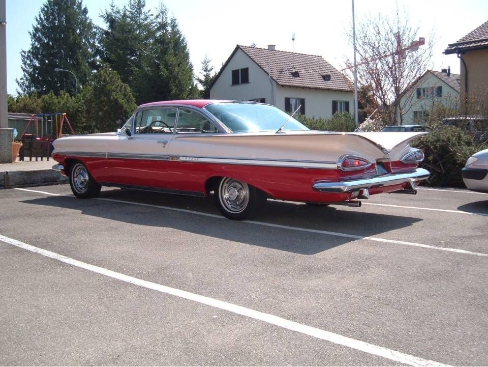 Ein zweifarbiger, rot-beiger Oldtimer steht auf einem Parkplatz; im Hintergrund sind Häuser und Bäume zu sehen.