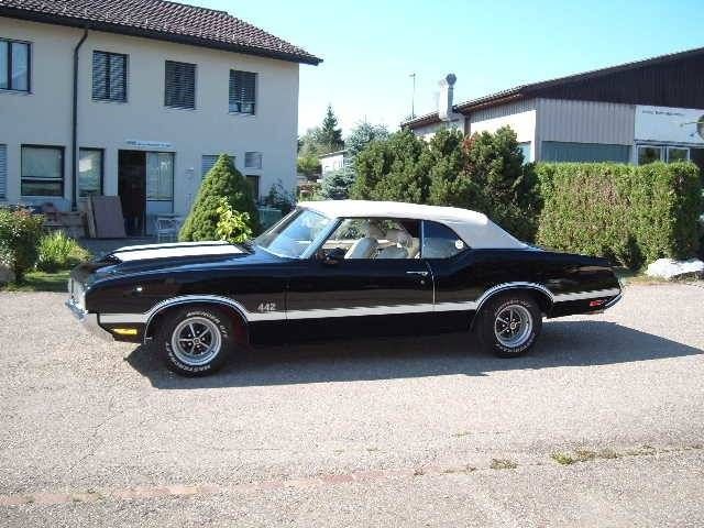 Schwarzes Oldsmobile 442 Cabriolet von 1970 mit weissem Verdeck und Rennstreifen, geparkt vor einem Gebäude.