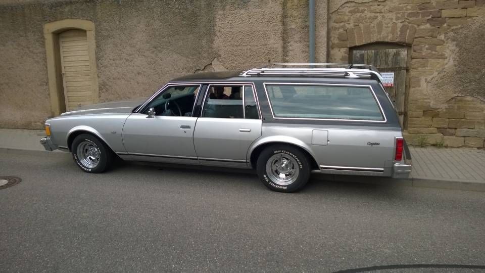 Ein silberner Kombi-Leichenwagen parkt auf einer Strasse; im Hintergrund ist ein Steingebäude zu sehen.