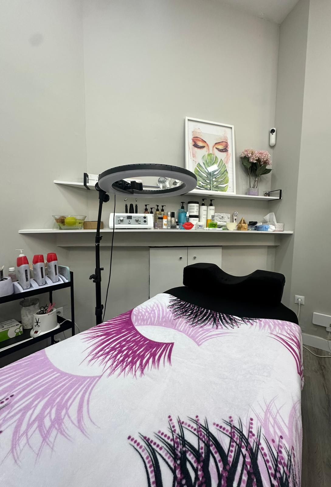 Una sala de tratamientos de belleza con camilla de masaje, anillo de luz y estantes con productos y obras de arte.