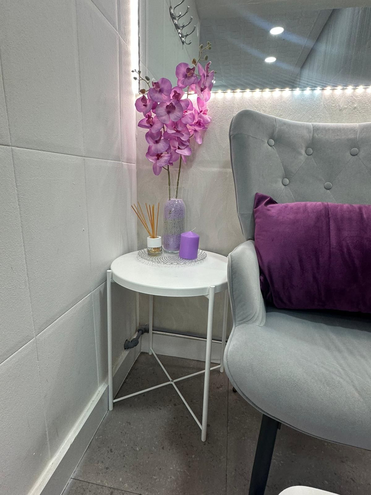 Una mesa auxiliar blanca con flores moradas, una vela y una silla con una almohada morada.