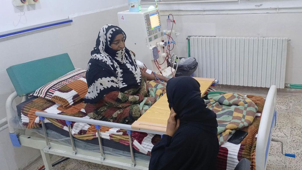 Eine Frau mit Kopftuch erhält in einem Krankenhausbett eine Dialyse. Eine andere Frau sitzt daneben. Medizinische Geräte sind sichtbar.