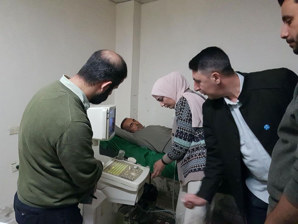 Menschen beobachten eine Ultraschalluntersuchung in einer medizinischen Einrichtung. Ein Patient liegt auf einer Liege, während ein Arzt das Gerät bedient.