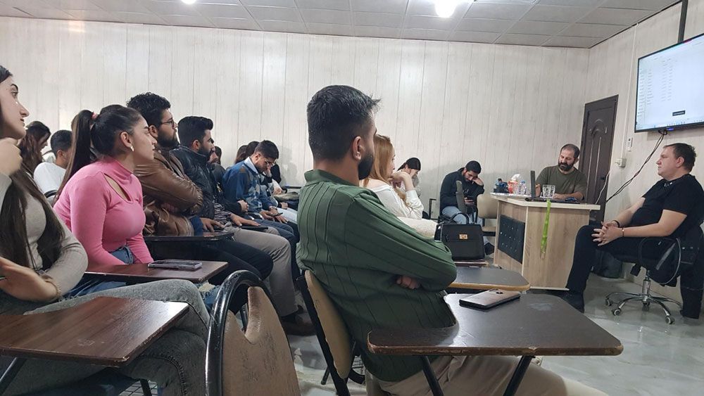 Ein Hörsaal mit Zuhörern, die zwei Rednern zuhören. Die Zuhörer sitzen an ihren Tischen und konzentrieren sich auf die Redner.