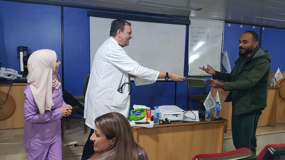 Personen in einer Präsentationssituation. Ein Mann im weißen Kittel überreicht einem Mann in einer grünen Jacke einen Gegenstand.