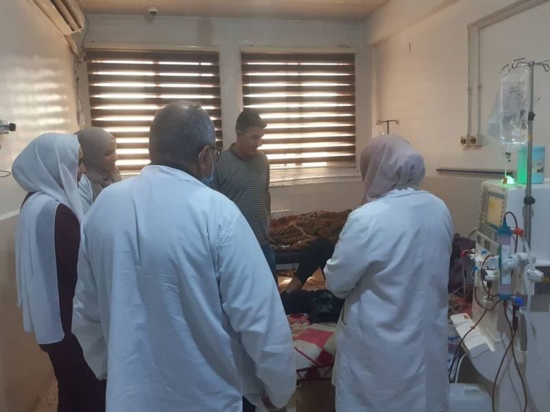 Medizinisches Personal in weißen Kitteln versammelt sich um ein Patientenbett in einem kleinen Krankenzimmer.