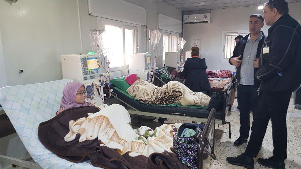 In einer medizinischen Einrichtung liegen Menschen in Betten. Zwei Männer stehen da und unterhalten sich. Ein Kind mit Kopftuch lächelt.