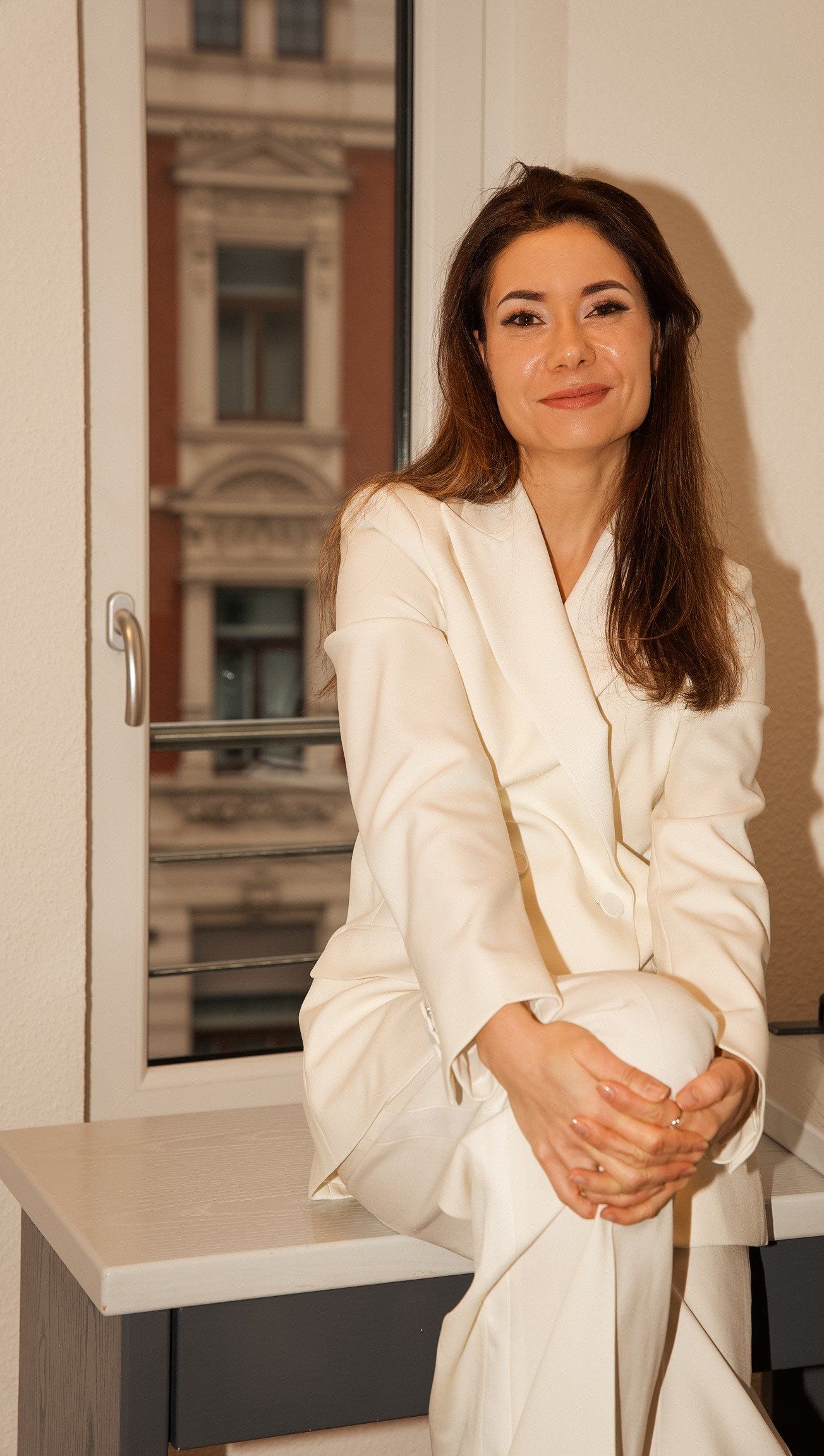 Eine Frau im weißen Anzug sitzt auf einem Fensterbrett.