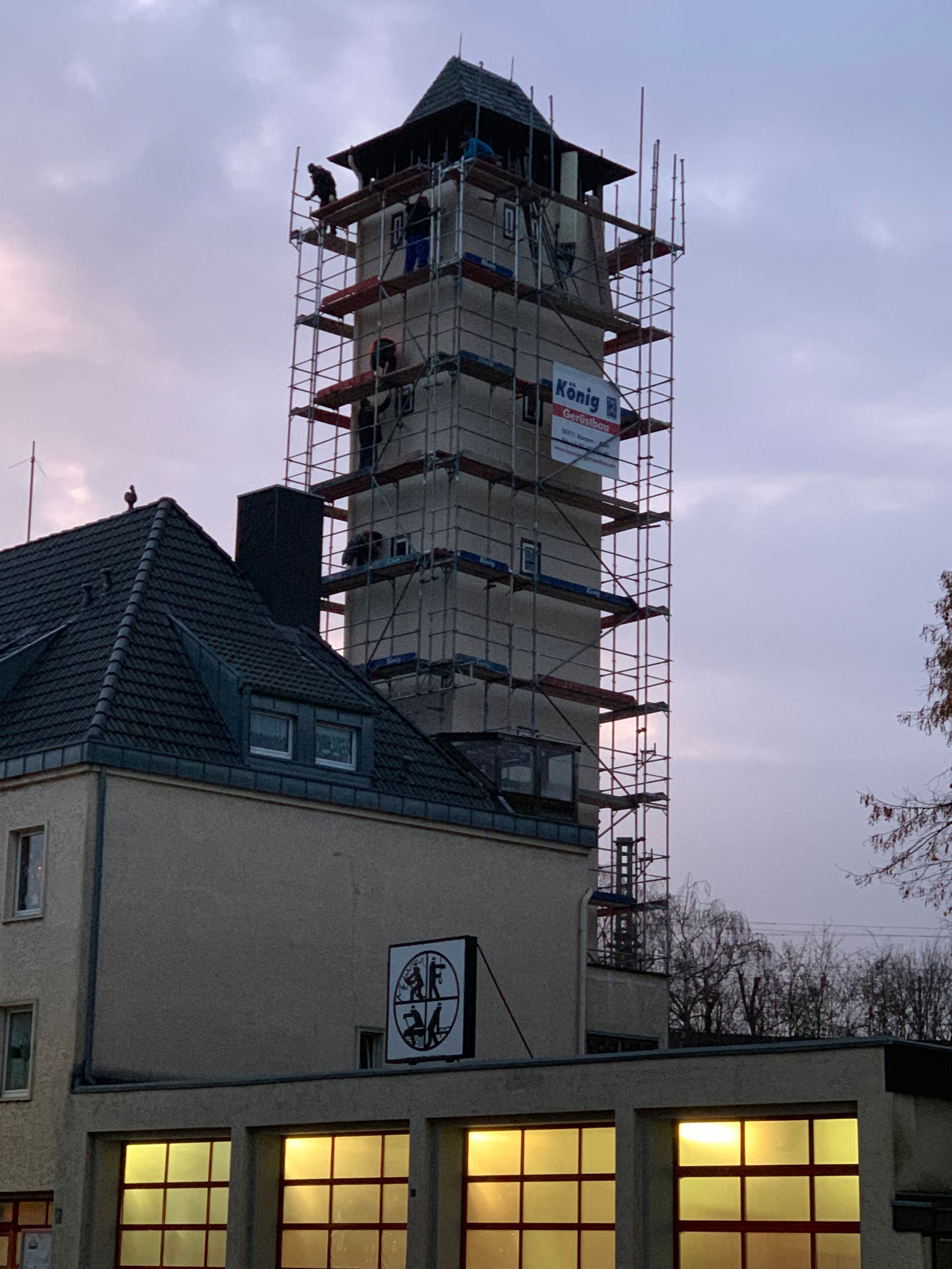Ein eingerüsteter Turm neben einem Gebäude mit erleuchteten Fenstern in der Abenddämmerung unter einem bewölkten Himmel