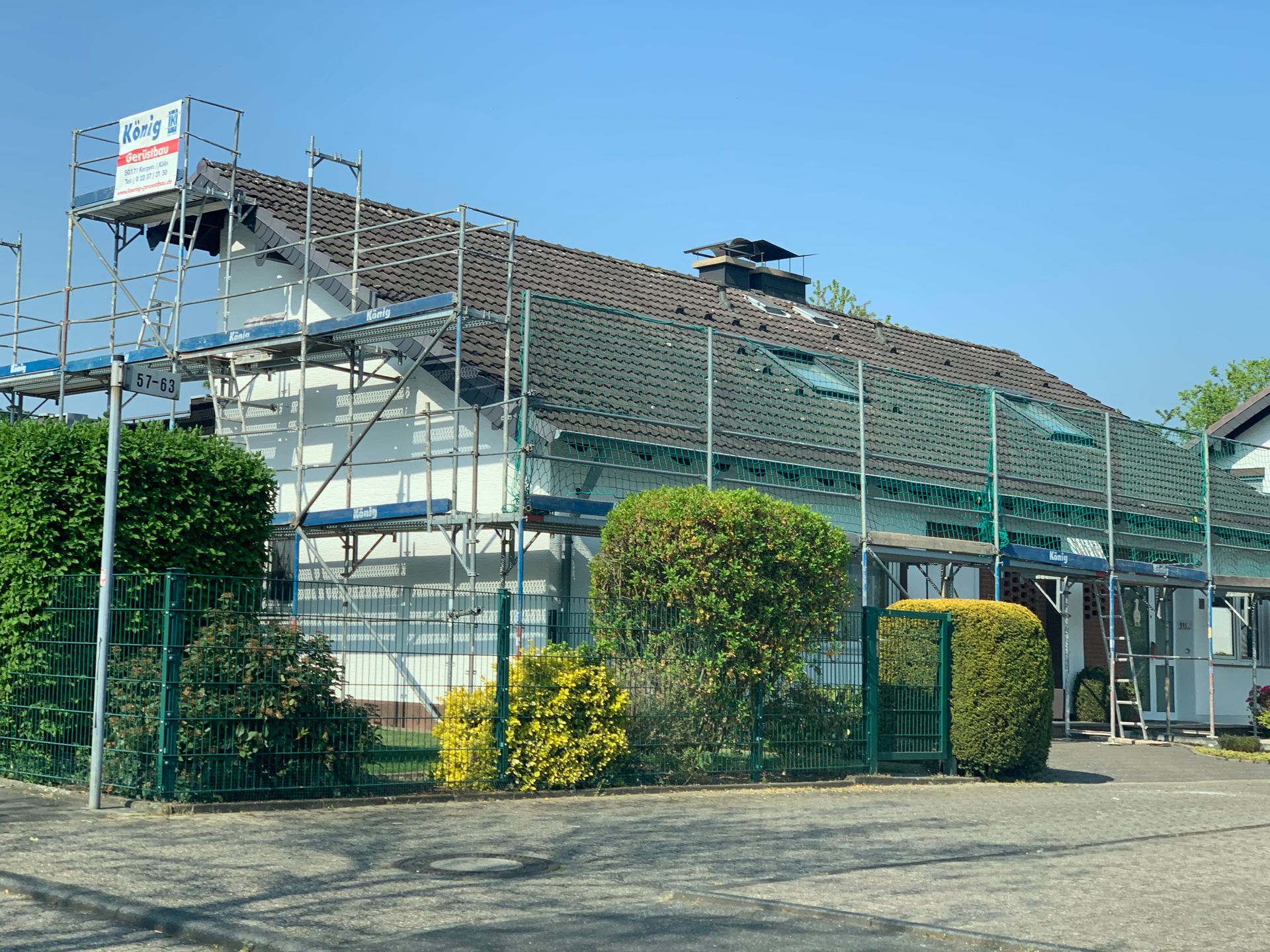 Solarpaneele auf dem Dach eines Gebäudes hinter einem Maschendrahtzaun und Sträuchern an einem sonnigen Tag
