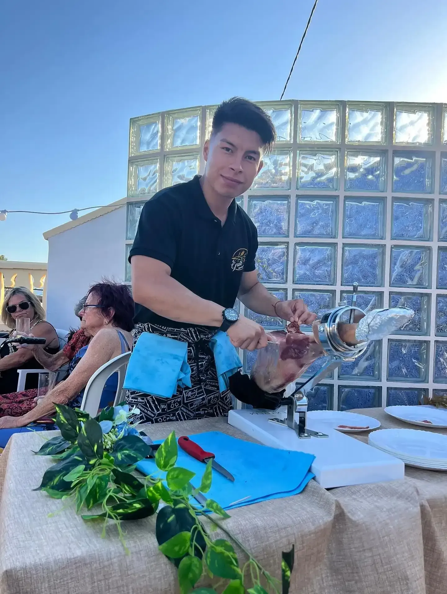 Hombre cortando jamón en un evento al aire libre, mirando a la cámara.