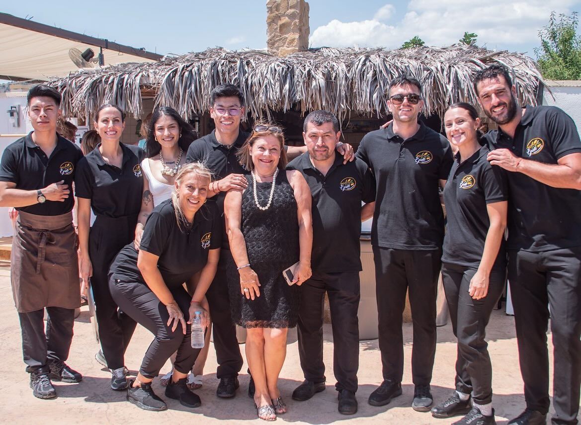 Grupo de personas, incluido personal, posando frente a un restaurante rústico con techo de paja, al aire libre.