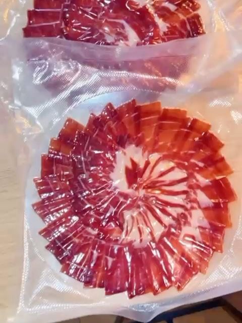 Jamón curado cortado en lonchas finas, dispuesto en espiral sobre un plato blanco, dentro de un paquete de plástico transparente.