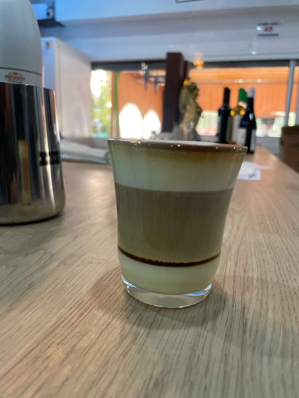 Vaso de bebida de café en capas sobre un mostrador de madera, con un fondo de cafetería borroso.