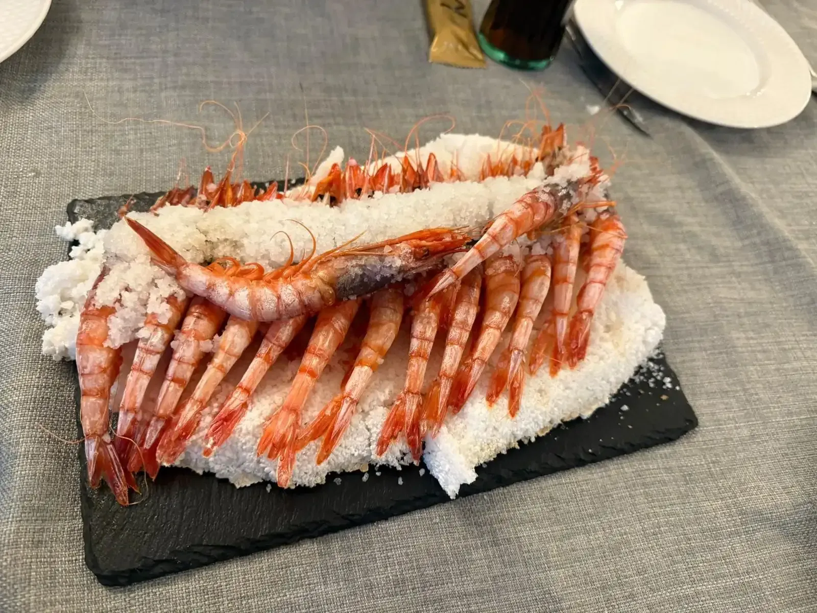 Camarones a la parrilla sobre un lecho de sal, servidos en un plato rectangular negro.