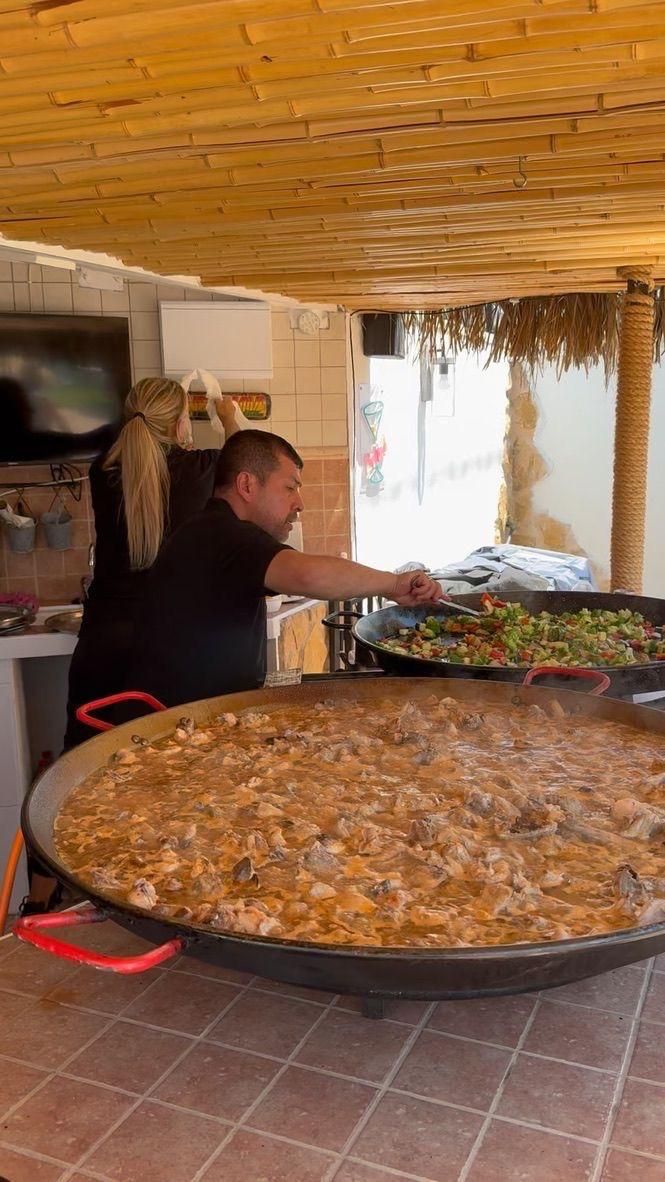 Hombre revolviendo una paella grande al aire libre con una sartén con mango rojo.