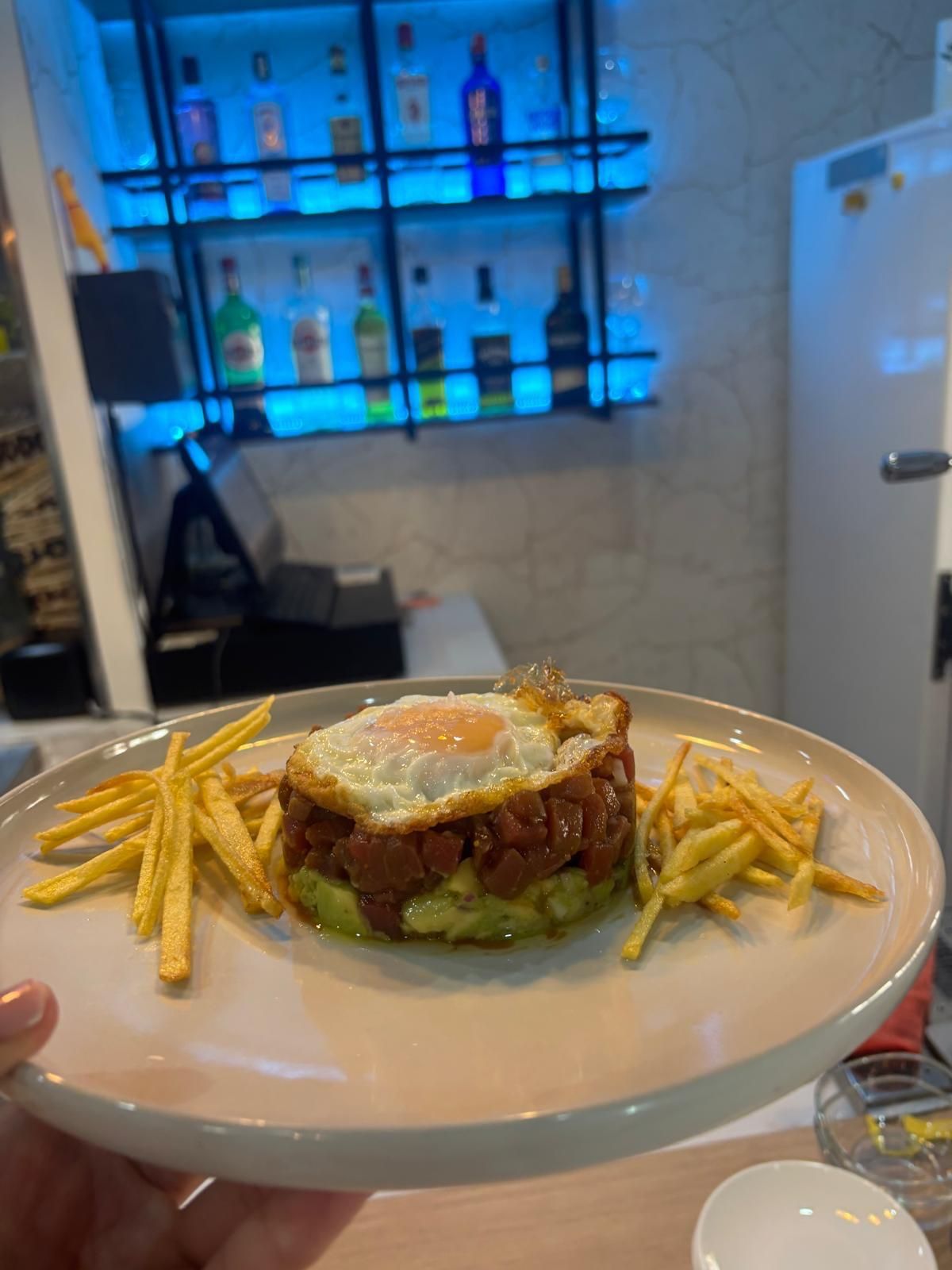 Plato con comida apilada: base de aguacate, carne, huevo frito, papas fritas, fondo de restaurante.