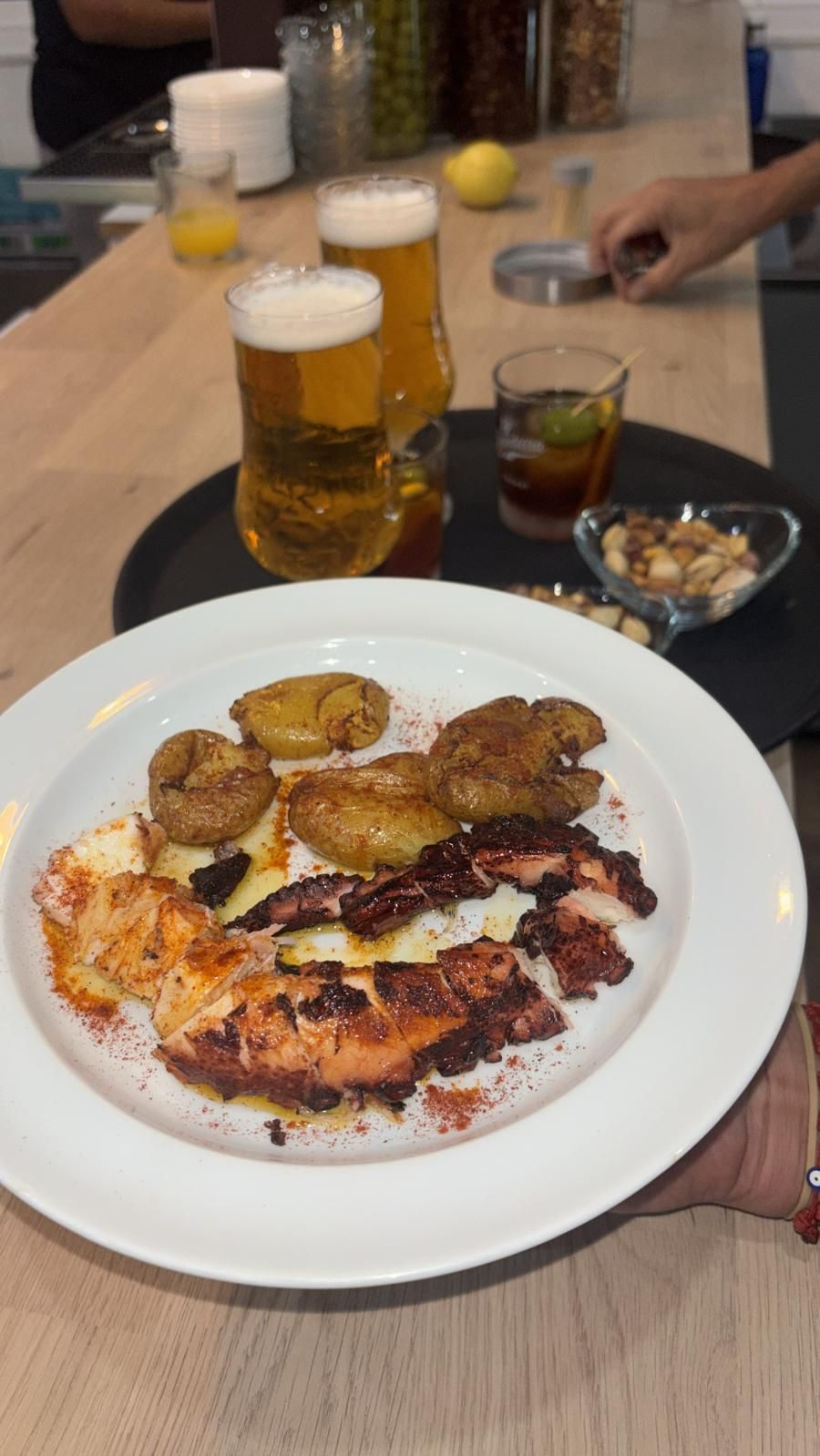 Plato de pulpo a la plancha, patatas y bebida en barra, con dos cervezas y frutos secos.