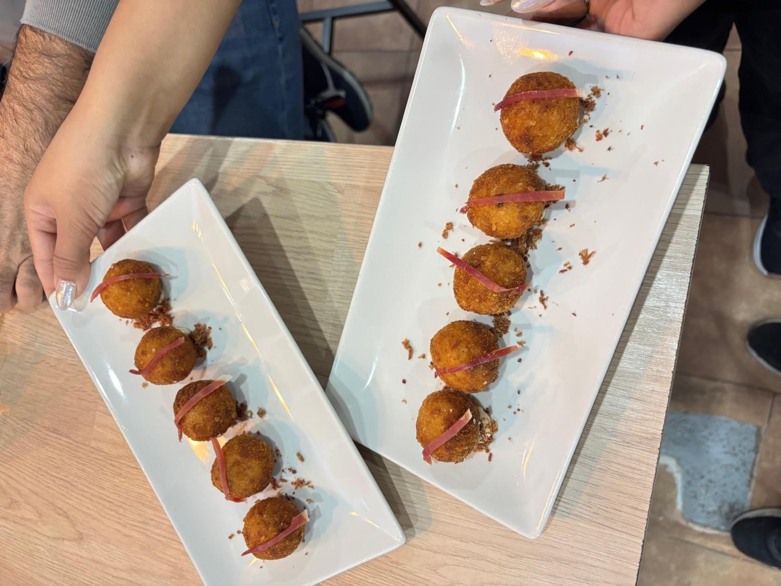 Se sostienen dos platos rectangulares blancos con comida frita, cubierta con tiras rojas y migas.
