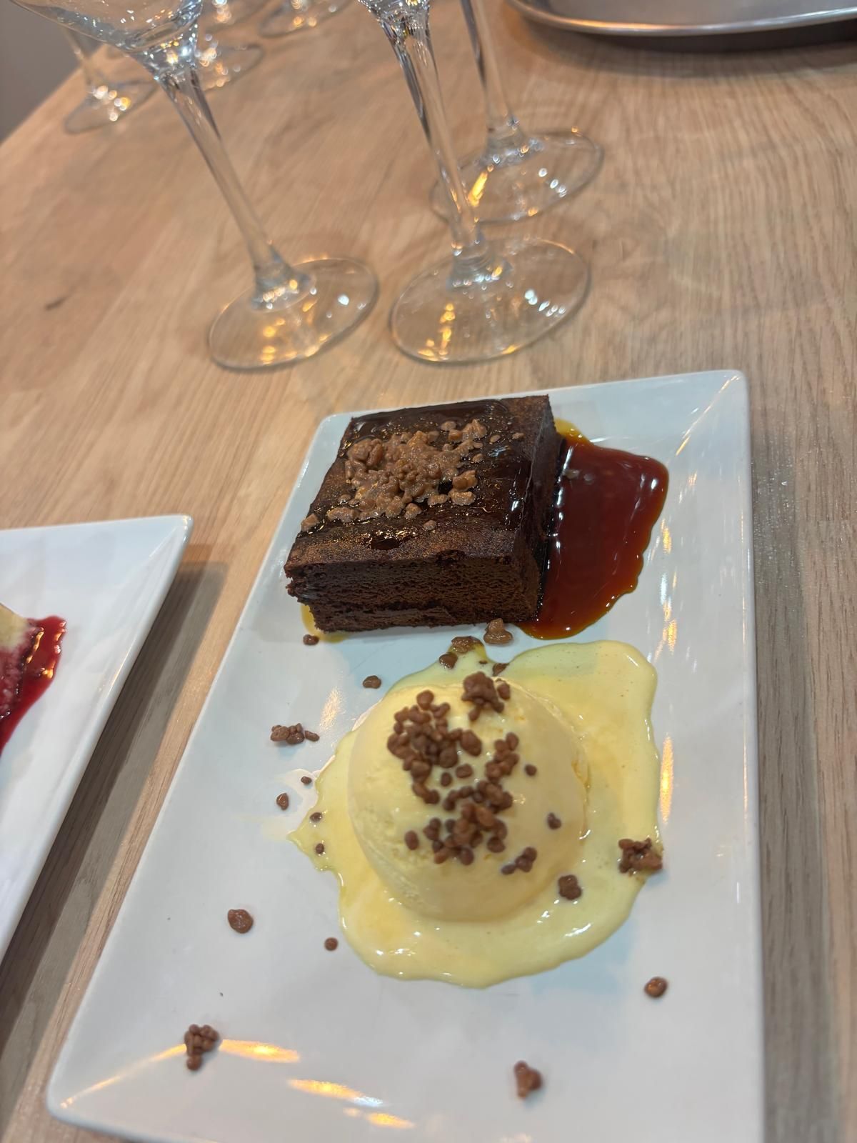 Brownie de chocolate con helado y salsa en un plato blanco; vasos en el fondo.