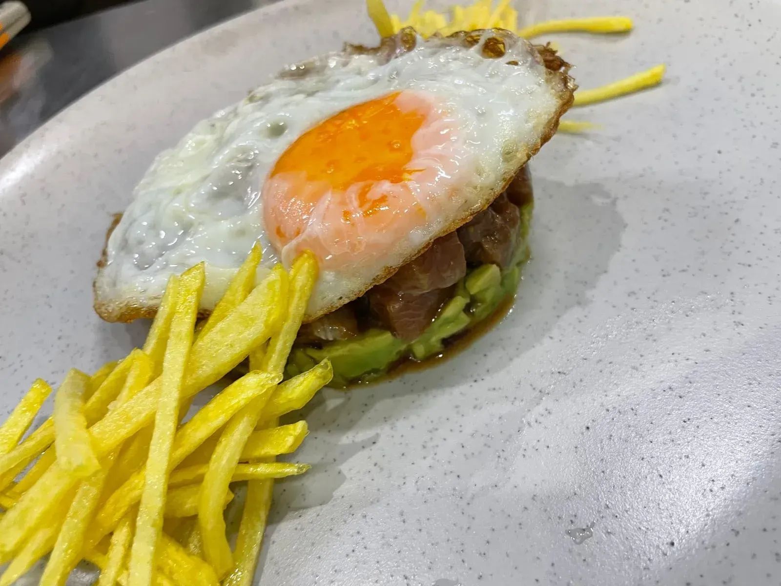 Plato con huevo frito sobre aguacate y atún, rodeado de patatas amarillas.