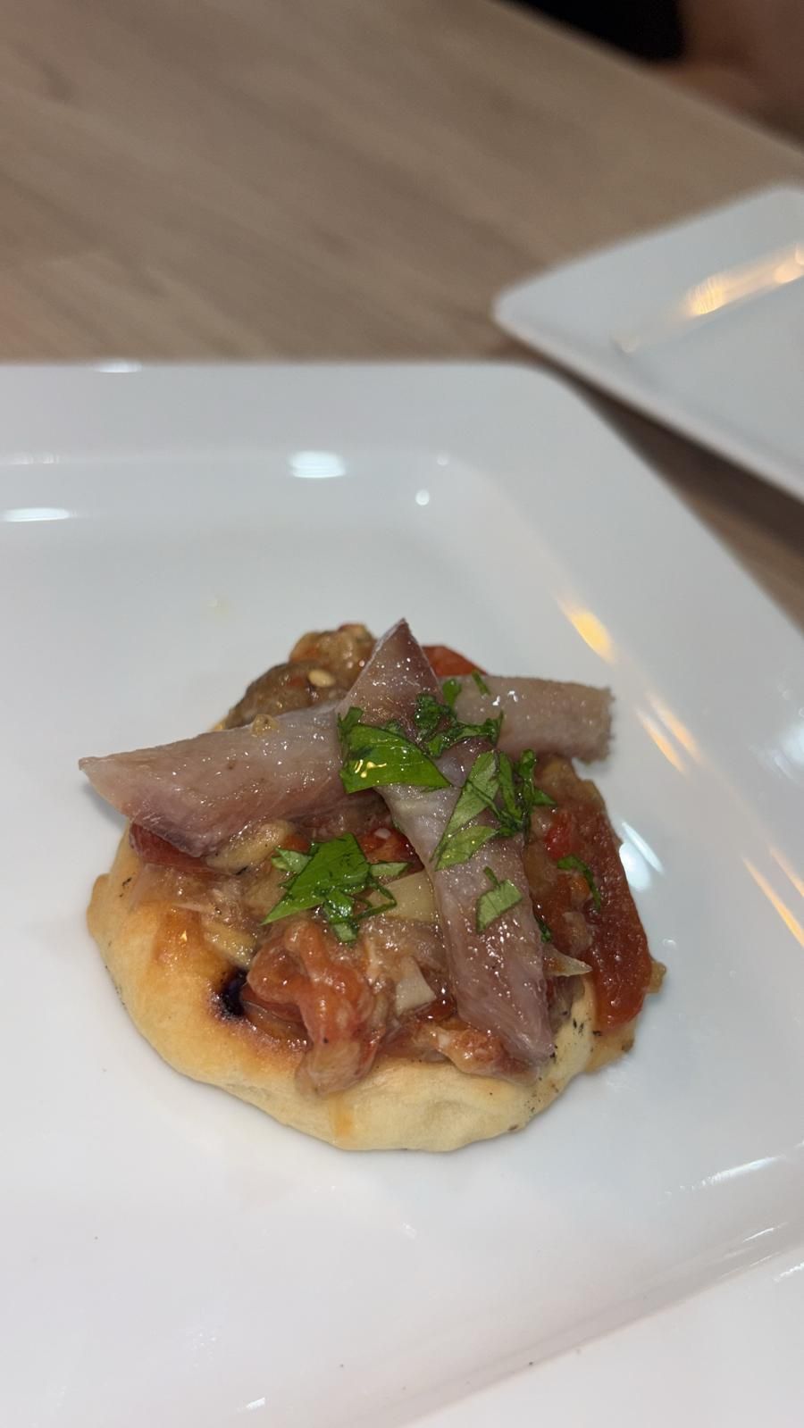 Pequeño aperitivo en plato blanco: pan tostado cubierto con tomates, anchoas y hierbas.