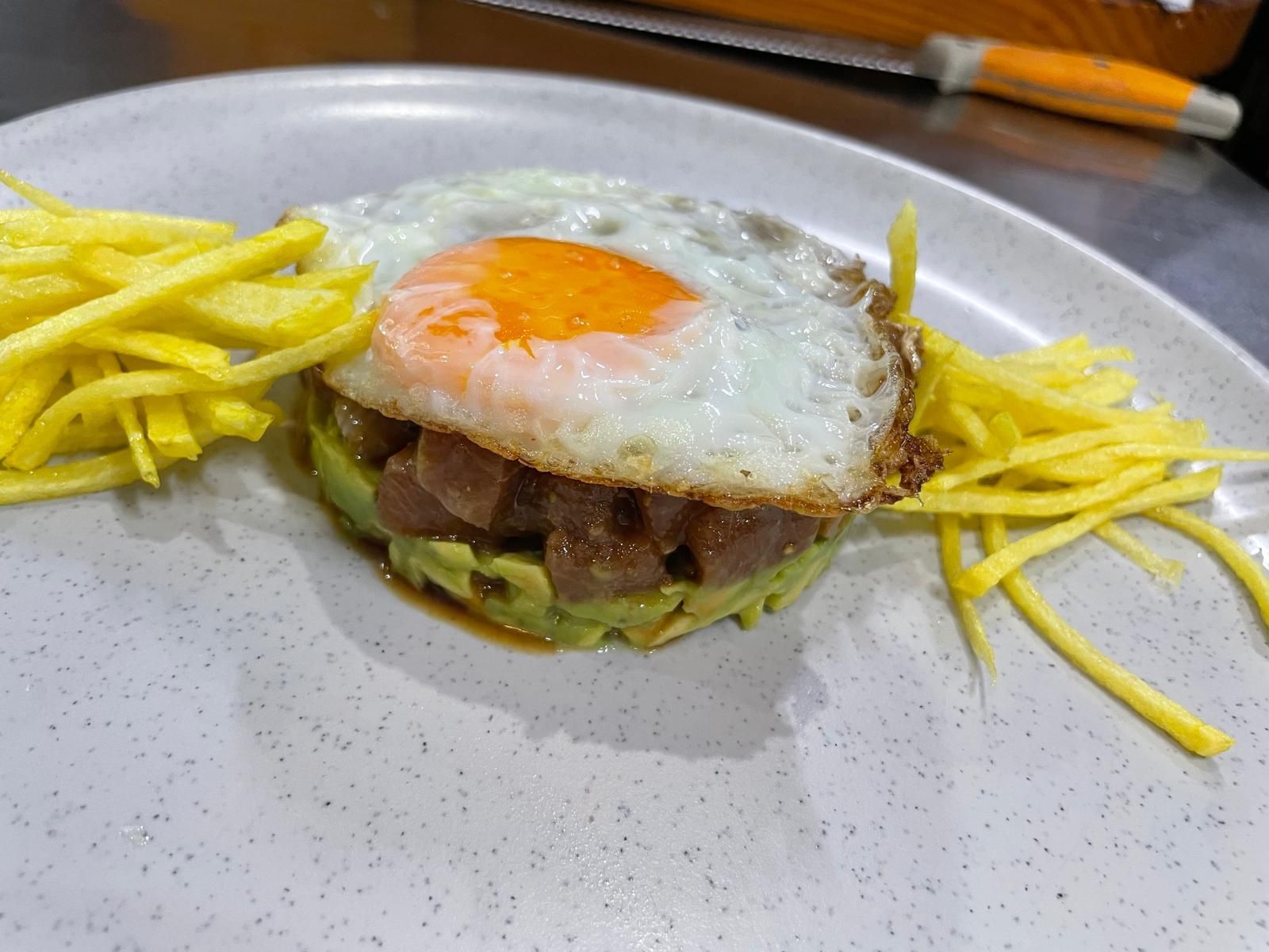 Tártaro de aguacate y atún cubierto con huevo frito, servido con juliana de mango en un plato.